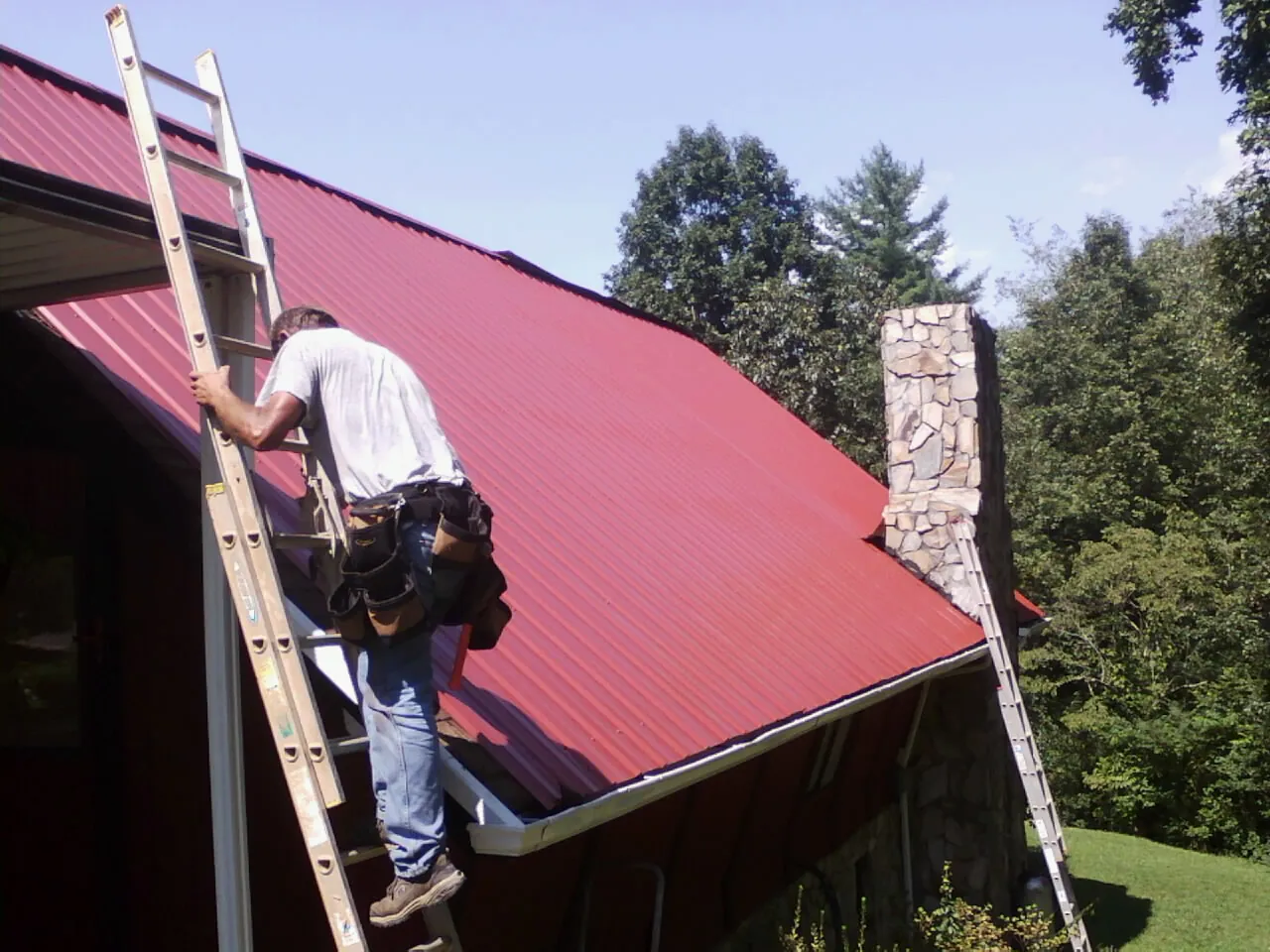 Roofer on a ladder beside a red metal roof and stone chimney — Soffit & Fascia Repair by Roofing Brothers in Green Village